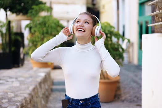 In an urban setting, a cheerful young woman with stylish headphones joyfully listens to music outdoors, embodying happiness, relaxation, and the exhilarating freedom that music brings