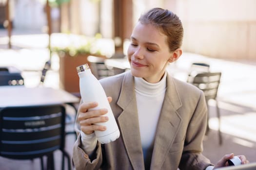 A smiling woman sitting at a caf table, admiring her stylish white water bottle in a vibrant outdoor setting