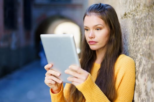 A young woman in a bright yellow sweater is intently using her tablet against a weathered stone wall in a lively urban setting, highlighting modern connectivity in our digital age