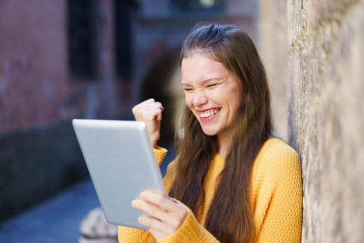 A young woman who is wearing a bright yellow sweater radiates joy as she smiles and celebrates, actively using a tablet, all set against a charming and historic backdrop that adds character