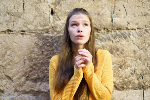 A young woman wearing a vibrant yellow sweater gazes thoughtfully upwards, her expression conveying a mixture of curiosity and hope, lost in her daydreams while leaning against a textured stone wall