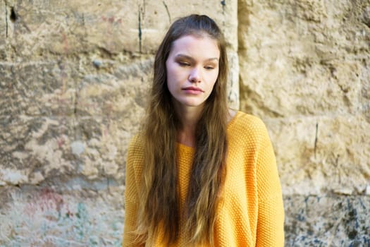 A young woman with long, flowing hair, dressed in a vibrant yellow sweater, stands in front of a textured stone wall, lost in deep contemplation, showcasing her beauty and calm demeanor