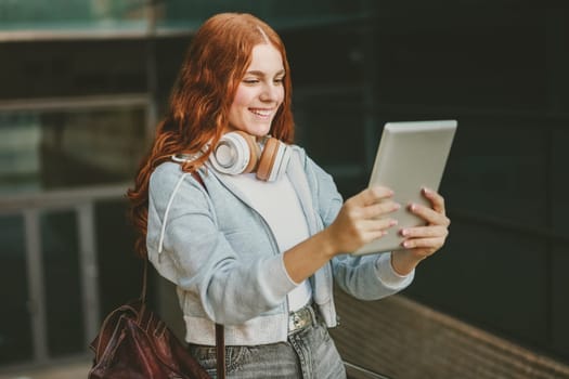 A cheerful and lively teenage girl with vibrant red hair is using a tablet to engage in a fun video call while wearing stylish headphones in a bustling urban environment, surrounded by city life