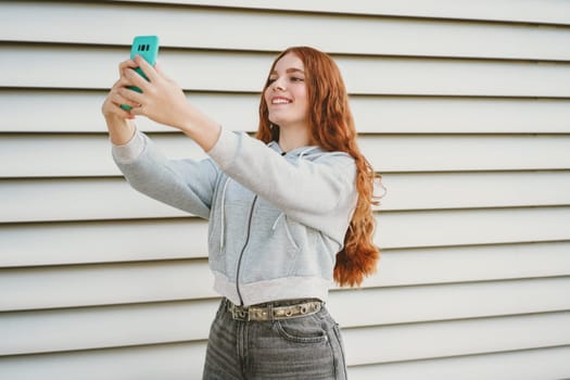 A joyful young woman is happily taking a selfie with her smartphone, showcasing her trendy casual style against a fascinating textured wall in a vibrant urban setting filled with life