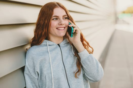 A cheerful young woman with long red hair is animatedly speaking on her phone, radiating positive energy while leaning against a stylish wall, ideal for lifestyle and communication themes