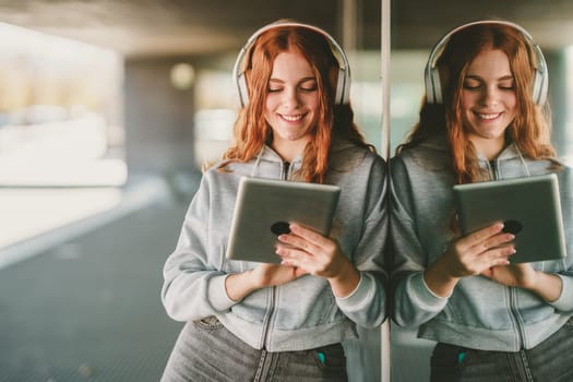 In a lively urban setting, a cheerful young woman with curly red hair finds joy through music, engaging with her tablet and wearing stylish headphones, fully immersed in her entertainment