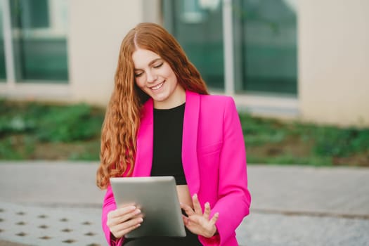 A cheerful young woman in a pink blazer is enjoying her outdoor leisure time. She holds a tablet while appreciating the fresh air, bright sunlight, and beautiful greenery around her