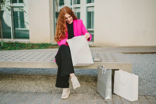 A stylish woman with beautiful curly hair is sitting comfortably on a bench in an urban setting, enthusiastically unpacking her shopping bags while showcasing the latest modern fashion trends