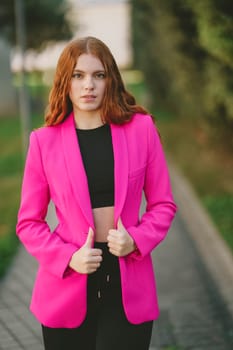 A stylish young woman confidently poses in a vibrant pink blazer and trendy black crop top, set against a beautifully blurred outdoor background that highlights her flair for modern fashion