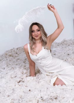 a very beautiful blonde is sitting in a white room with feathers flying in it. the girl has long hair. she is wearing a white silk dress. on top she is holding a large feather in her hand