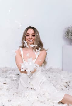 a very beautiful blonde is sitting in a white room with feathers flying in it. the girl has long hair styled in a wave. she is wearing a white silk dress. she is holding feathers in her hands