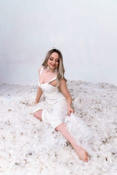 a very beautiful blonde is sitting in a white room with feathers flying in it.the girl has long hair. she is wearing a white silk dress. she is holding a large feather in her hand and touching her leg