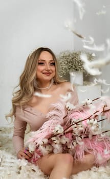 young beautiful blonde girl among feathers. she sits among white feathers. they fly around her. in her hands she holds pink big feathers and branches of cotton