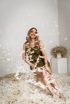 a very beautiful blonde sits in a white room in which feathers are flying. the girl has long hair styled in a wave. dressed in a dark silk dress. white fluff is flying in the air