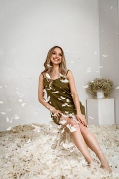 a very beautiful blonde is sitting in a white room with feathers flying . she is wearing a dark silk dress. white fluff is flying in the air. a lot of feathers are flying in front of her face
