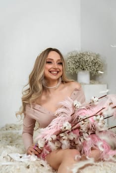 young beautiful blonde girl among feathers. she sits among white feathers and smiles. feathers fly through the air. in her hands she holds pink big feathers and branches of cotton