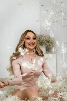 young beautiful blonde girl among feathers. she sits among white feathers and smiles. feathers fly in the air. she threw feathers into the air with her hands and is happy about it