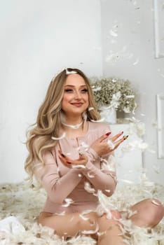 young beautiful blonde girl among feathers. she sits among white feathers and smiles. feathers fly in the air. girl crossed her arms. she has long blonde hair, she has pearls on her neck