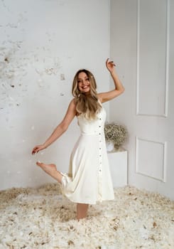 young beautiful girl stands in a room where feathers are flying. feathers are flying through the air in a white room. blonde girl with long hair, dressed in a long white dress. she is dancing