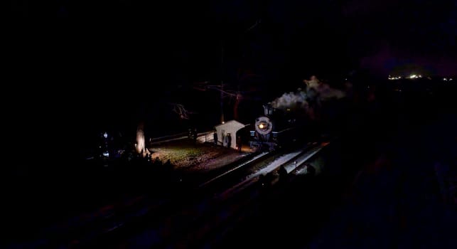 A vintage steam locomotive emits a plume of smoke as it pauses at a quaint station during the night. Soft lights from the station enhance the serene atmosphere, while stars twinkle above.