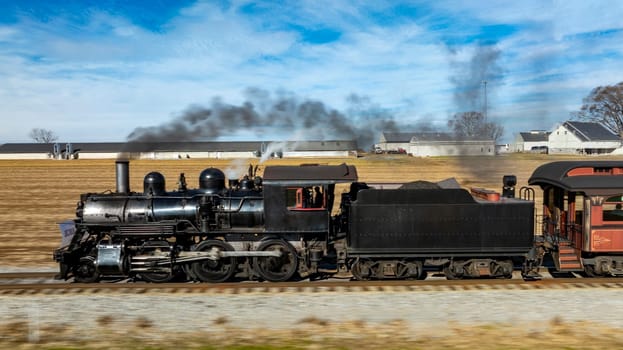 A vintage steam locomotive powers along the tracks, sending plumes of smoke into the blue sky. Surrounding fields and farmhouses enhance the picturesque countryside setting.