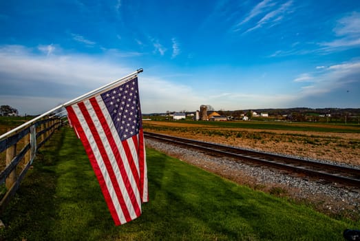 The American flag stands proudly next to railway tracks in a tranquil rural setting during sunset. Fields stretch out in the background under a vibrant sky.