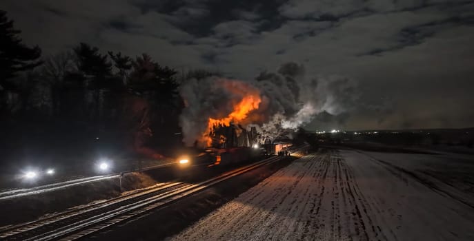 A train car is on fire next to snowy fields under a moonlit sky. Smoke billows into the air as emergency vehicles shine lights on the scene, creating an urgent atmosphere.