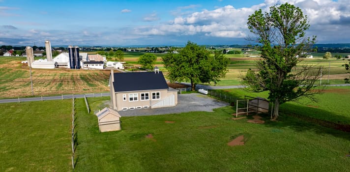 An Amish one room school house sits on a gravel area, surrounded by expansive green fields, barns, and distant structures under a bright blue sky.