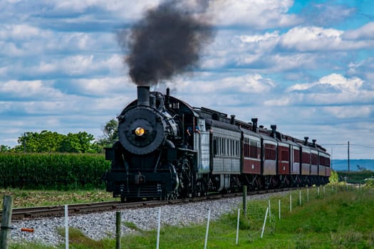 A vintage steam train puffs black smoke as it moves along the railway, surrounded by lush green cornfields and under a bright blue sky with fluffy clouds, creating a picturesque countryside view.