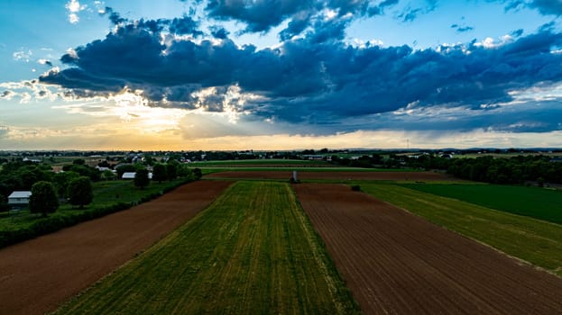 A vibrant sunset casts warm colors over expansive farmland, with lush green fields and patches of clouds creating a picturesque sky. The serene landscape reflects the tranquility of rural life.