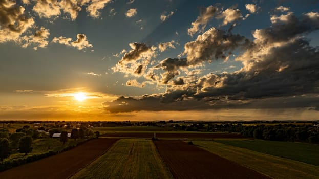 A vibrant sunset casts warm tones across expansive fields, framed by dramatic clouds. The landscape features greenery and farm structures under a colorful sky, showcasing a tranquil evening.