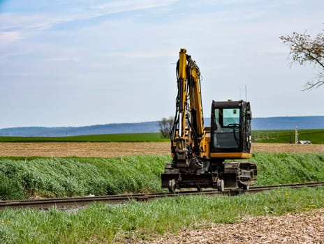 A yellow excavator maneuvers alongside a railway track in a flat rural area. The machine is performing maintenance work amidst green grass and fields under a clear sky.