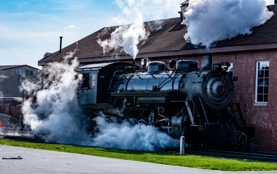 The powerful steam locomotive prepares for departure at a historic railway station. Billowing clouds of steam rise amidst bright sunlight, creating a captivating display of industrial nostalgia.
