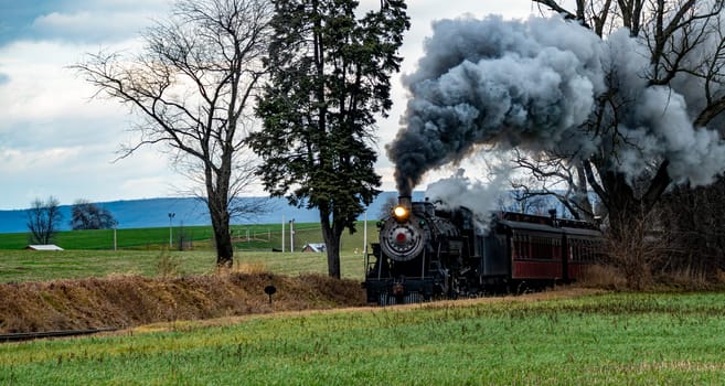 A vintage steam train chugs along railway tracks in a vast rural area, emitting thick plumes of smoke against a backdrop of dark clouds. Nearby fields exhibit golden crops.