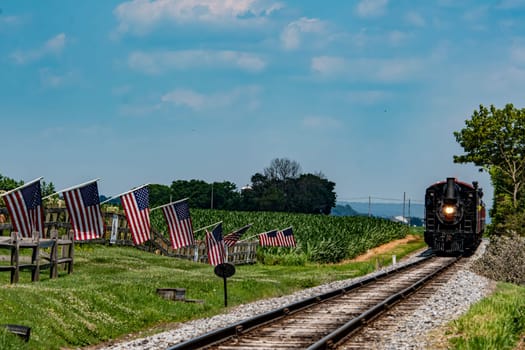 A classic steam train moves along the tracks in lush green countryside. American flags line the pathway, adding color to the bright blue sky and warm sunlight.
