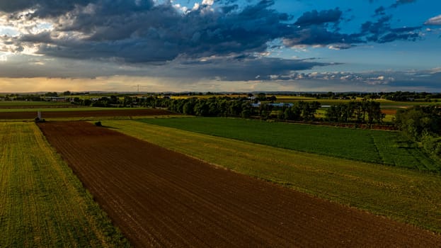 As the sun sets in a vibrant sky, vast agricultural fields stretch across the landscape, dotted with trees and farm structures, creating a serene and picturesque rural scene.