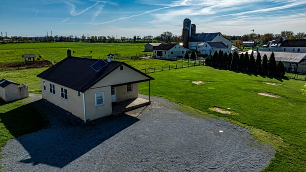 An Amish one room school house sits on a gravel area, surrounded by expansive green fields, barns, and distant structures under a bright blue sky.