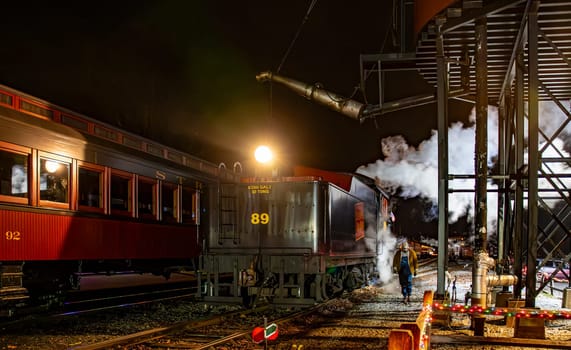 Strasburg, Pennsylvania, USA December 7, 2024 - Vintage steam locomotive is illuminated at night as engineers takes water on the locomotive. The surrounding area features a passenger car.