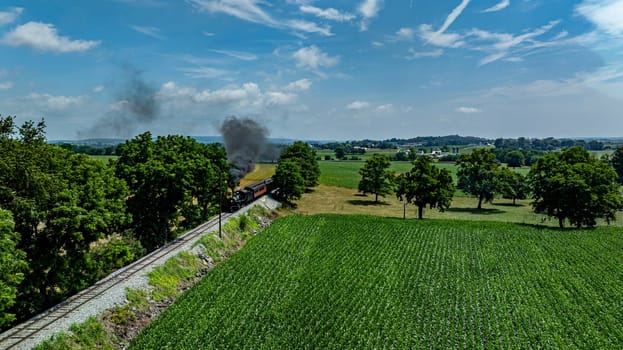 A steam locomotive chugs along tracks surrounded by lush green fields and trees on a bright day. The landscape features a clear blue sky with wispy clouds, enhancing the scenic view.