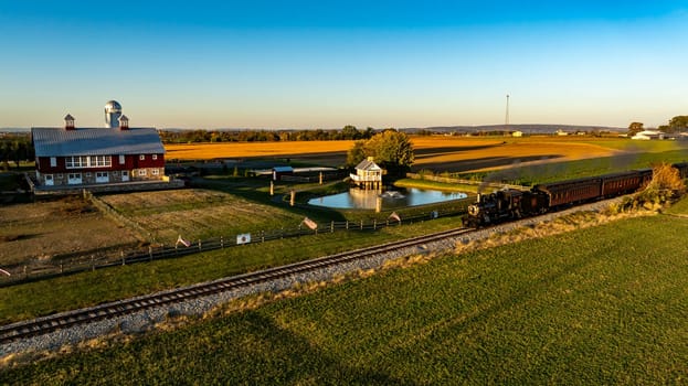 Ronks, Pennsylvania, USA, October 20 2024 - Historic steam train moves along the tracks, passing a picturesque red barn expansive farmland. The sun sets, casting warm golden hues across the landscape,