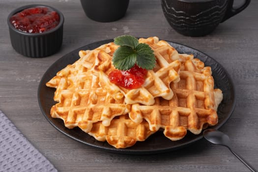 Delicious waffles topped with strawberry jam on a black plate with a grey wooden background.