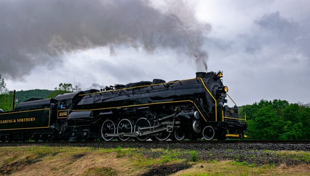 Ringgold, Pennsylvania, USA, May 28, 2022 - A classic steam locomotive chugs along a track, releasing clouds of steam as it journeys through verdant fields under a gray sky, showcasing timeless travel.