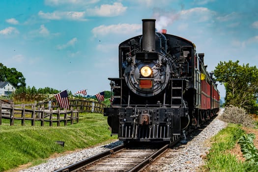 A vintage steam train moves along its tracks in a picturesque countryside, with blue skies overhead and flags lining the route. The scene captures an old-fashioned travel experience.