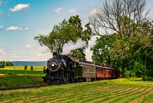 A steam train chugs along railway tracks bordered by vibrant green fields. The sun shines down, illuminating the historic engine and colorful carriages amid a tranquil landscape.