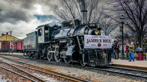 Strasburg, Pennsylvania, USA, December 31, 2024 - A steam locomotive, adorned with a farewell banner, stands at a bustling railway station. Rail enthusiasts gather to witness its last journey, creating a nostalgic atmosphere.