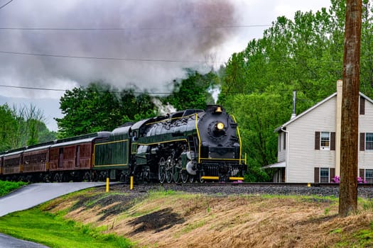 A vintage steam train, black with yellow trim, makes its way along a rural track under gray skies. Houses and lush trees line the route, emphasizing the peaceful countryside setting.