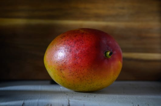A ripe mango sits gracefully on a weathered wooden backdrop, reflecting warm hues of yellow and red under soft light.