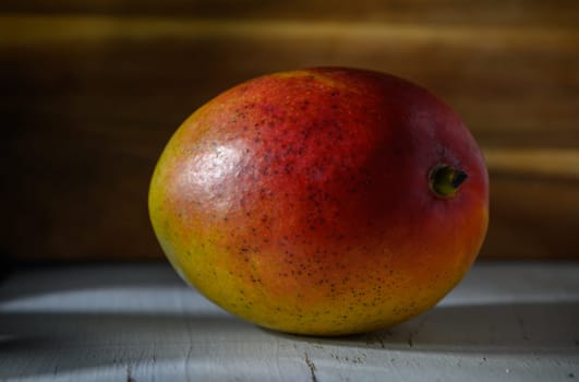 A ripe mango with bright red and yellow hues sits on a weathered surface, showcasing its juicy appeal in warm light.