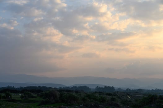 Golden light of sunset illuminates a tranquil countryside with hills and softly swirling clouds on the horizon.