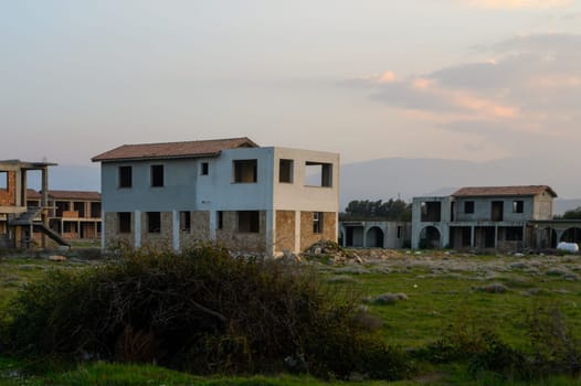 Ruins of unfinished constructions stand silently as the sun sets, casting shadows across the overgrown landscape.